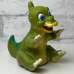 May include: A vintage green dinosaur figurine with a yellow beak, blue eyes, and white accents on its hands and feet. The dinosaur is sitting with its arms raised. The figurine is a collectible toy.