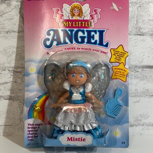 May include: A blue and silver My Little Angel doll with wings and a rainbow background. The doll has a blue dress and a pink sash. The doll's name is Mistie. The packaging includes the text "Magical Glow in the Dark" and "Gently shake and watch the magic in the wings!"