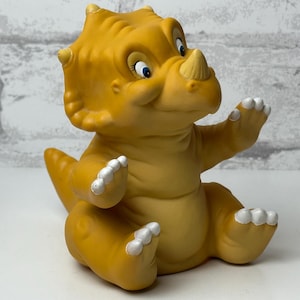 May include: A yellow dinosaur figurine with a friendly expression. The figurine has a horn, small horns, and white claws. The dinosaur is sitting with its arms raised. The background is a white brick wall.