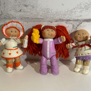 Puede incluir: Tres muñecas Cabbage Patch Kids, una con un vestido rojo y blanco, otra con un pijama morado y un oso de peluche amarillo, y otra con un vestido morado y blanco y una muñeca rosa.