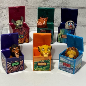 Puede incluir: Una colección de seis figuras de personajes de El Rey León en cajas con bisagras de colores. Cada caja presenta un personaje diferente, como Simba, Mufasa, Scar, Ed, Rafiki y Pumbaa. Las cajas están decoradas con ilustraciones temáticas y nombres de personajes.