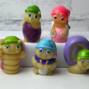 Op de afbeelding: Een verzameling van zes kleine, grillige beeldjes. De figuren zijn onder meer een rups, een slak en andere wezens, allemaal met cartoonachtige kenmerken en hoeden. Het kleurenpalet omvat geel, paars, roze en blauw.