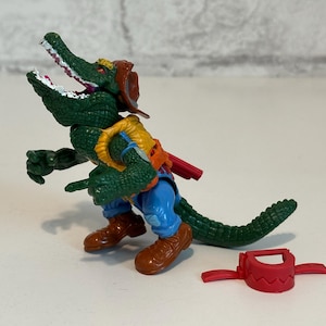 Puede incluir: Una figura de acción de cocodrilo verde que lleva un sombrero de vaquero marrón, pantalones vaqueros azules y un chaleco amarillo. La figura tiene una funda roja y una pistola. La figura está de pie sobre una superficie blanca.