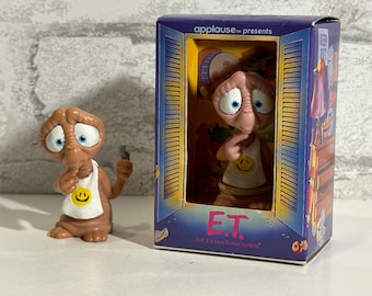 May include: A brown and tan E.T. figurine with a yellow smiley face on a white shirt. The figurine is in a plastic box with the words "Applause presents E.T. The Extra-Terrestrial" on the front.