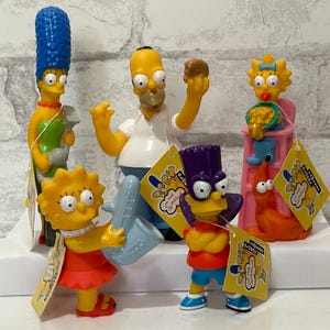 May include: A set of six Simpsons character figurines, including Homer, Marge, Bart, Lisa, Maggie, and a dog. The figurines are made of plastic and are brightly coloured. Each figurine has a small tag attached to it with the Simpsons logo and the character's name.