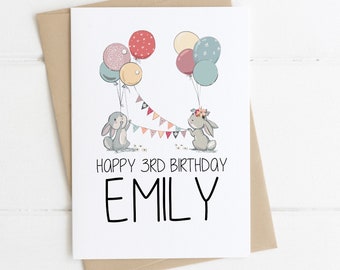 Puede incluir: Una tarjeta de cumpleaños con dos conejos sosteniendo una cuerda de banderas coloridas. Los conejos sostienen globos. La tarjeta dice "Feliz 3er cumpleaños Emily".