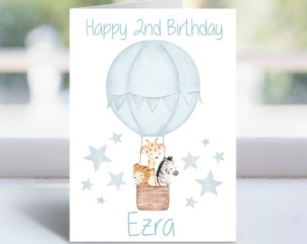 May include: A blue and white hot air balloon with a giraffe, zebra, and lion inside. The balloon is surrounded by white stars. The card says "Happy 2nd Birthday Ezra".