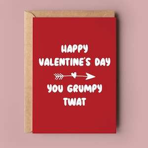 May include: A red Valentine's Day card with the text "Happy Valentine's Day You Grumpy Twat" in white. The card is inside a brown envelope, set against a pink background.