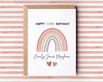 Op de afbeelding: Een witte kaart met een roze, beige en grijze regenboogillustratie. De tekst "Happy Third Birthday" staat boven de regenboog en de naam "Emily Jones Mayhem" staat onder de regenboog. Twee rode harten staan onder de naam.