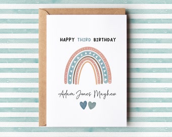 Op de afbeelding: Een kleurrijke regenboog-verjaardagskaart met de tekst "Happy Third Birthday" en de naam "Adam Jones Mayhew" in handschrift. Twee blauwe hartjes staan onder de naam.