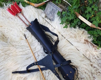 Op de afbeelding: Een zwarte leren koker met een schouderriem en een houten pijl. De koker is gevuld met rode en witte gevederde pijlen. De koker ligt op een wit, pluche oppervlak.