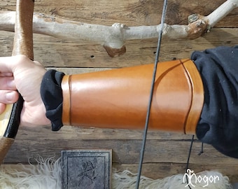 May include: A brown leather arm guard with a black strap. The arm guard is worn on the left arm and is being held in place by a hand holding a bow.