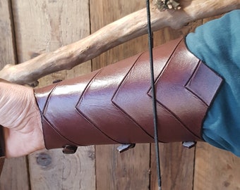 May include: A brown leather arm guard with a chevron pattern. The arm guard is worn on the left arm and is secured with straps. The arm guard is being held by a person who is also holding a bow.