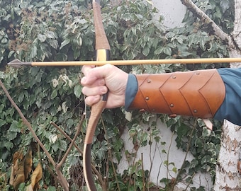 May include: A person holding a wooden bow and arrow, wearing a brown leather arm guard with decorative stitching. The bow is made of wood and has a black string. The arrow is made of wood and has a metal tip. The person is wearing a blue shirt and is standing in front of a green leafy background.
