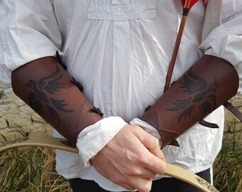 May include: A person wearing brown leather arm guards with a bird design. The arm guards are secured with straps and buckles. The person is holding a bow and arrow.
