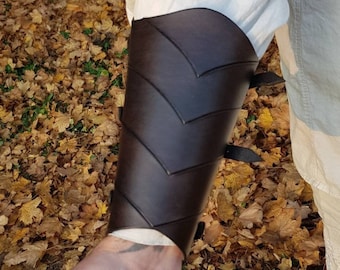 May include: Brown leather arm guard with a chevron pattern and straps. The arm guard is worn on the left arm and is secured with straps.