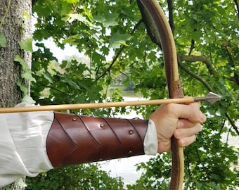 May include: A brown leather arm guard with metal accents, worn on the left arm of a person holding a bow and arrow. The person is outdoors, with a green tree and a body of water in the background.