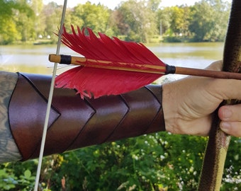May include: A brown leather arm guard with a chevron pattern, worn on a person's arm. A red-feathered arrow is held in the person's hand, with the arrow shaft resting on the arm guard.