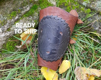 May include: A brown leather arm guard with a black embossed design of a stylized animal head. The arm guard has a strap with a buckle.