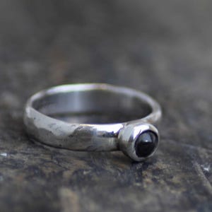 May include: A silver ring featuring a hammered, textured band and a single, small, round, polished black stone set in a smooth, circular bezel setting. The metal has a reflective, metallic finish with visible indentations along the band.