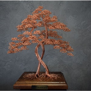 Op de afbeelding: Een koperdraadsculptuur van een boom met verweven stammen en een volle kruin. De boom staat op een houten voet met een kleine plaquette. De sculptuur staat tegen een donkergrijze, getextureerde achtergrond.