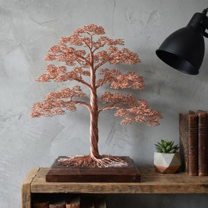 Op de afbeelding: Een koperdraad boomsculptuur met een gedraaide stam en gedetailleerde takken, gemonteerd op een donkere houten basis. De boom staat op een rustieke houten plank met een kleine vetplant en vintage boeken. Een zwarte lamp is in de buurt geplaatst.