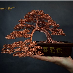 May include: A copper wire bonsai tree sculpture in a dark wooden rectangular pot. The tree has a curved trunk and detailed branches with small leaves. The pot has Chinese characters. The background is dark.