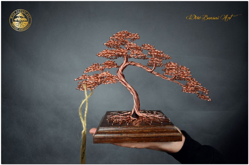 May include: A copper wire bonsai tree sculpture with a brown wooden base. The tree is in a classic bonsai style with a thick trunk and many branches. The sculpture is on a wooden base and is being held by a hand.