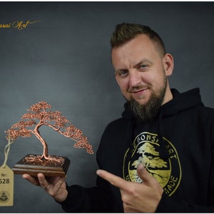 May include: A person holding a copper wire bonsai tree sculpture on a wooden base. The sculpture is numbered 528. The person is wearing a black hoodie with a gold logo that says "Wire Bonsai Art" and a tree design.