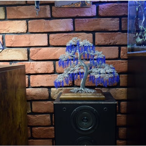 May include: A wire sculpture of a weeping willow tree with blue beads hanging from the branches. The sculpture is displayed on a wooden base and sits on top of a black speaker.