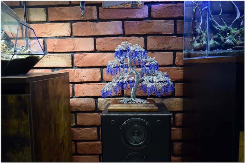May include: A wire sculpture of a weeping willow tree with blue beads hanging from the branches. The sculpture is displayed on a wooden base and sits on top of a black speaker.