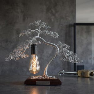 May include: A table lamp featuring a silver-colored wire sculpture shaped like a bonsai tree. The tree has a curved trunk and multiple branches with fine wire foliage. A single Edison-style light bulb hangs from a branch above a wooden base.