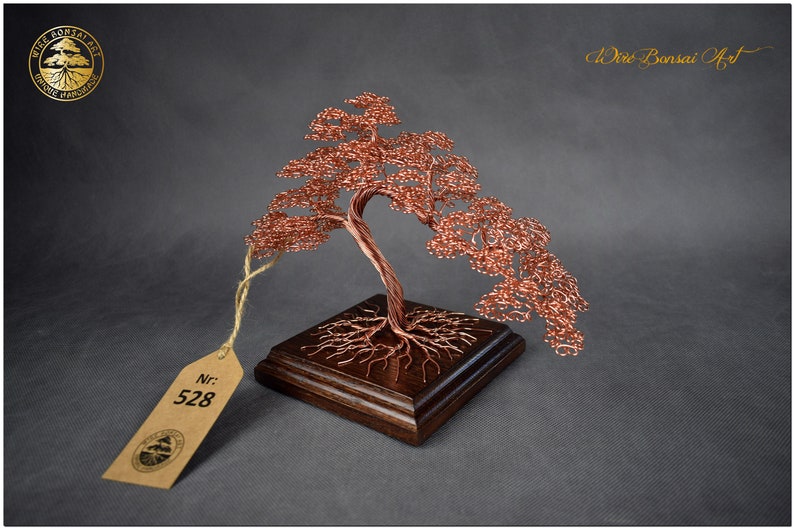 May include: A copper wire bonsai tree sculpture with a brown wooden base. The tree is shaped like a weeping willow and has a tag with the number 528 on it.