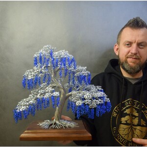 May include: A silver wire sculpture of a weeping willow tree with blue beads for leaves. The tree is on a wooden base and is being held by a person wearing a black hoodie with a logo that says "Wire Bonsai Art Unique Handmade".