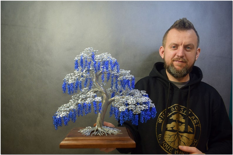 May include: A silver wire sculpture of a weeping willow tree with blue beads for leaves. The tree is on a wooden base and is being held by a person wearing a black hoodie with a logo that says "Wire Bonsai Art Unique Handmade".