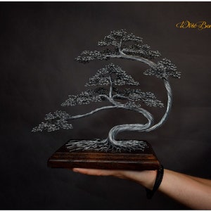 K&ouml;nnte beinhalten: Handgefertigte Draht-Bonsai-Baumskulptur. Der Baum hat ein detailliertes Silberdrahtdesign mit einem dunklen Holzsockel. Die Skulptur wird in einer Hand vor dunklem Hintergrund gehalten. Das Logo lautet "MIKE BONSAI ART UNIQUE HANDMADE".