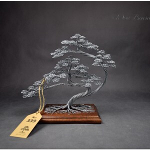 K&ouml;nnte beinhalten: Silberdraht-Bonsai-Baumskulptur mit verdrehtem Stamm und geschichtetem Laub, montiert auf einem Holzsockel. Die Skulptur hat ein Etikett mit der Nummer 339. Der Hintergrund ist dunkelgrau.