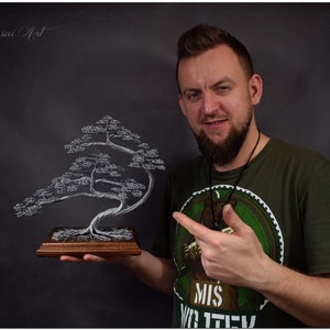 K&ouml;nnte beinhalten: Handgefertigte Draht-Bonsai-Baumskulptur. Der Baum ist silberfarben und auf einem rechteckigen Holzsockel montiert. Die Skulptur wird von einer Person vor einem dunklen Hintergrund gehalten. Der Text "Wire Bonsai Art" ist sichtbar.