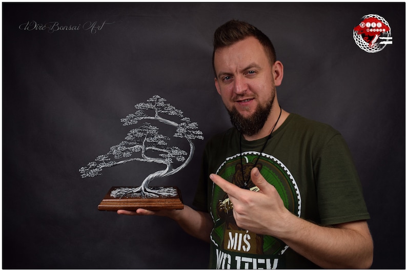 K&ouml;nnte beinhalten: Handgefertigte Draht-Bonsai-Baumskulptur. Der Baum ist silberfarben und auf einem rechteckigen Holzsockel montiert. Die Skulptur wird von einer Person vor einem dunklen Hintergrund gehalten. Der Text "Wire Bonsai Art" ist sichtbar.