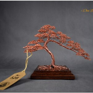 May include: A copper wire bonsai tree sculpture with a brown wooden base. The tree has a twisted trunk and branches with small copper wire leaves. The sculpture is labeled with the number "528".