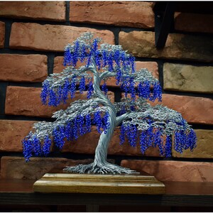 May include: A wire sculpture of a weeping willow tree with blue beads representing the leaves. The tree is displayed on a wooden base against a brick wall.