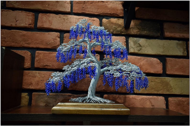 May include: A wire sculpture of a weeping willow tree with blue beads representing the leaves. The tree is displayed on a wooden base against a brick wall.