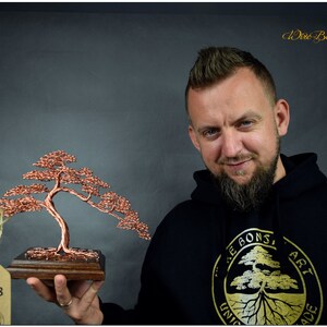May include: A copper wire bonsai tree sculpture on a wooden base. The sculpture is being held by a person wearing a black hoodie with a gold logo that says "Wire Bonsai Art Unique Handmade". A tag with the number "Nr: 528" is attached to the sculpture.