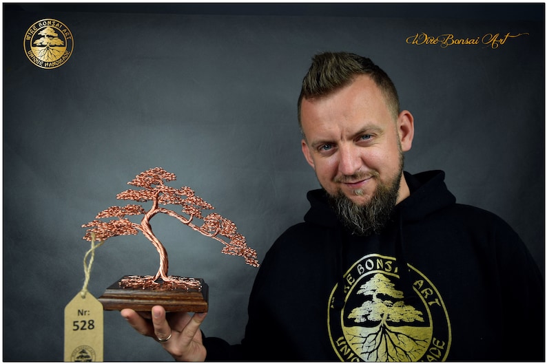 May include: A copper wire bonsai tree sculpture on a wooden base. The sculpture is being held by a person wearing a black hoodie with a gold logo that says "Wire Bonsai Art Unique Handmade". A tag with the number "Nr: 528" is attached to the sculpture.