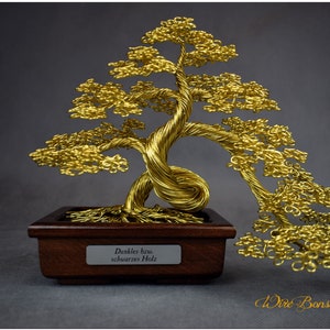 Op de afbeelding: Een gouden draadbonsai-boombeeldhouwwerk op een donkerbruine houten basis. De basis heeft een label met de tekst "Dunkles Holz", wat in het Nederlands "Donker Hout" betekent.