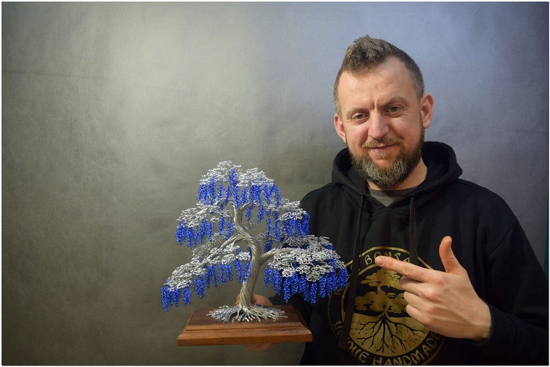 May include: A silver wire sculpture of a weeping willow tree with blue beads for leaves. The tree is on a wooden base and is being held by a person wearing a black hoodie with a tree logo.