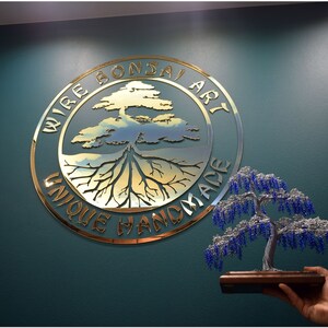 May include: A gold metal sign with the text "Wire Bonsai Art Unique Handmade" in a circle with a stylised tree and roots inside. A small blue and silver beaded bonsai tree is held in a hand.