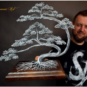K&ouml;nnte beinhalten: Handgefertigte Draht-Bonsai-Kunstskulptur. Der Baum hat ein detailliertes Design aus silbernem Draht mit einem Holzsockel. Die Skulptur hat eine nat&uuml;rliche Baumform mit komplizierten &Auml;sten und Bl&auml;ttern. Der Sockel ist rechteckig und aus dunklem Holz.