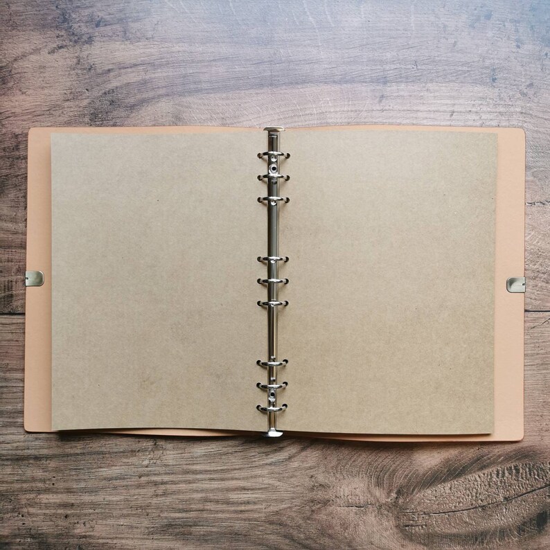 May include: An open, tan-colored notebook with a metal binder, lying on a wooden surface. The notebook's pages are blank, offering space for notes or sketches. The cover is a lighter tan color.