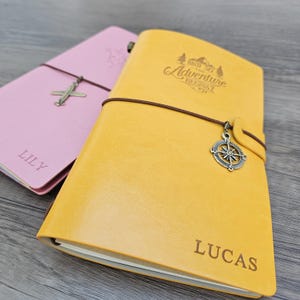 Può includere: Due diari in pelle, uno rosa e uno giallo, con chiusure elastiche marroni. Il diario rosa ha un ciondolo a forma di aeroplano e il nome "LILY" inciso. Il diario giallo presenta un ciondolo a forma di bussola e la frase "AND SO THE ADVENTURE BEGINS" con il nome "LUCAS" inciso.